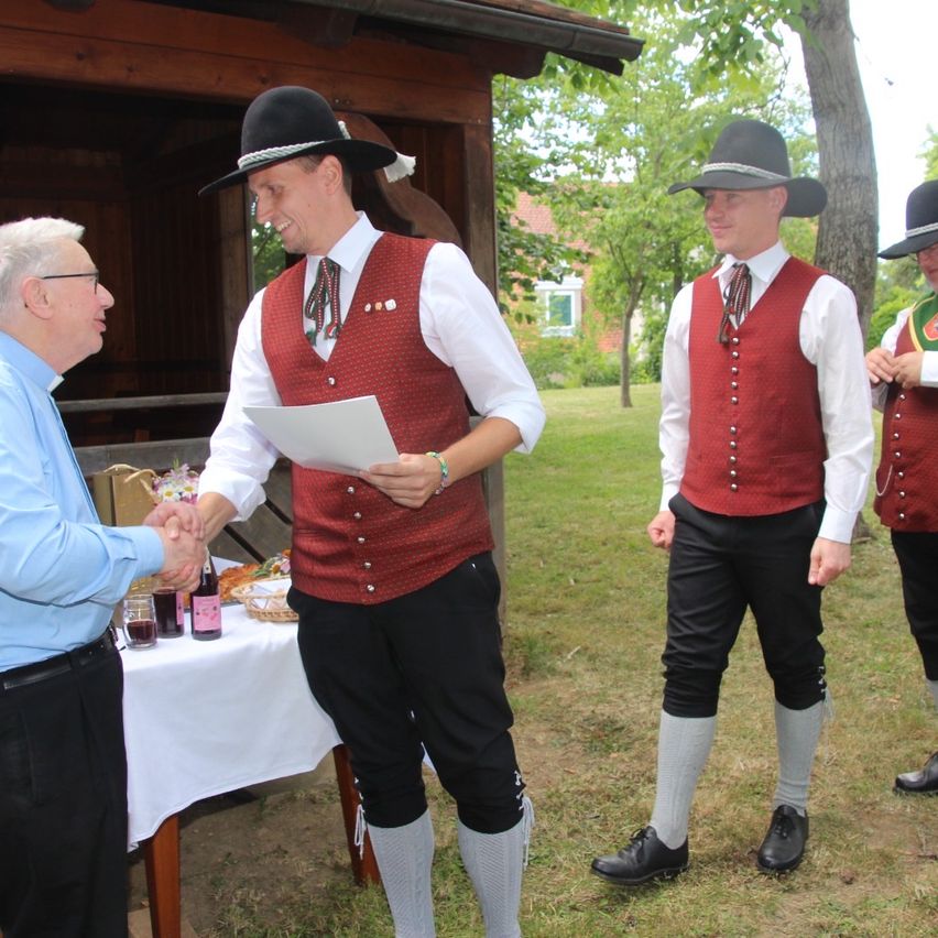 Three men in traditional Bavarian attire stand outdoors, one holding a paper, another holding a handshake, and a table with drinks in the background.