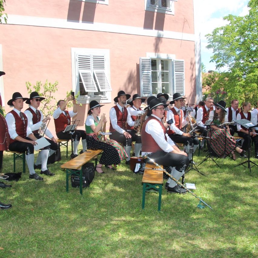 A group of musicians dressed in traditional attire are seated on benches in a grassy area, possibly preparing for a performance. Behind them is a building with white shutters.