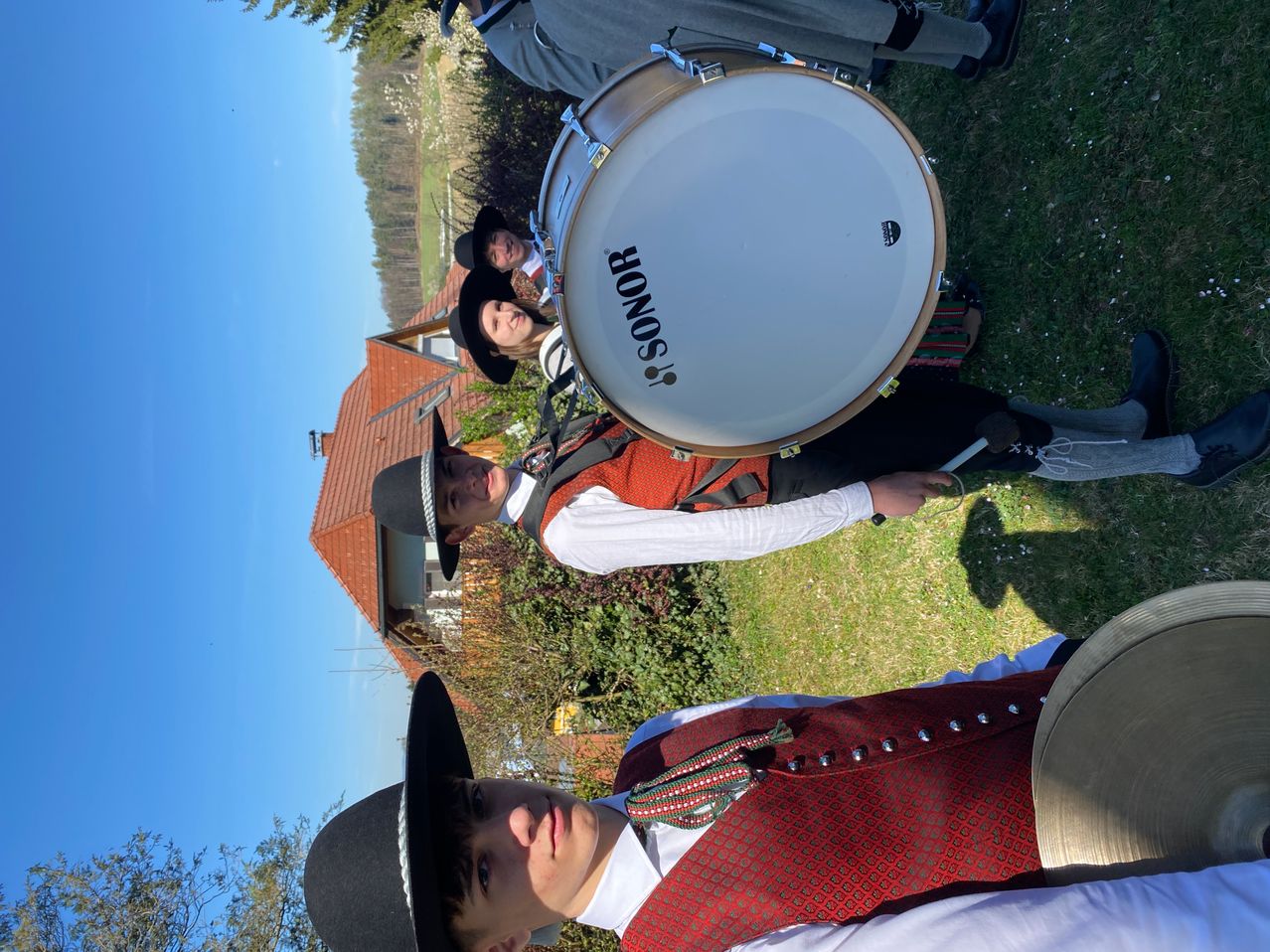 Three individuals dressed in traditional attire, one holding a drum and the other two looking on, with a house in the background.
