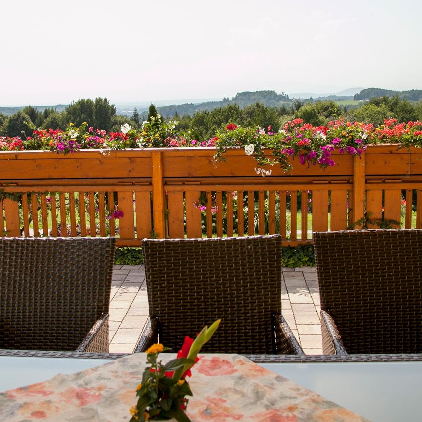 Ein Balkon mit einem hölzernen Geländer, mit Blick auf eine Landschaft voller verschiedener Blumen und Grün. Drei braune Weidenstühle sind aufgestellt, mit einer Blumentischdekoration im Vordergrund.