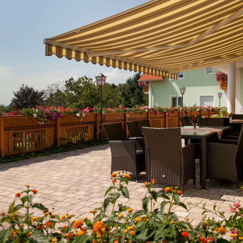 Ein Garten mit einem Terrassendach, Korbstühlen und einem Tisch. Die Terrasse hat einen Ziegelboden und eine Hecke mit Blumenkästen. Ein Gebäude mit rotem Dach und Glasfenstern steht hinter der Hecke.