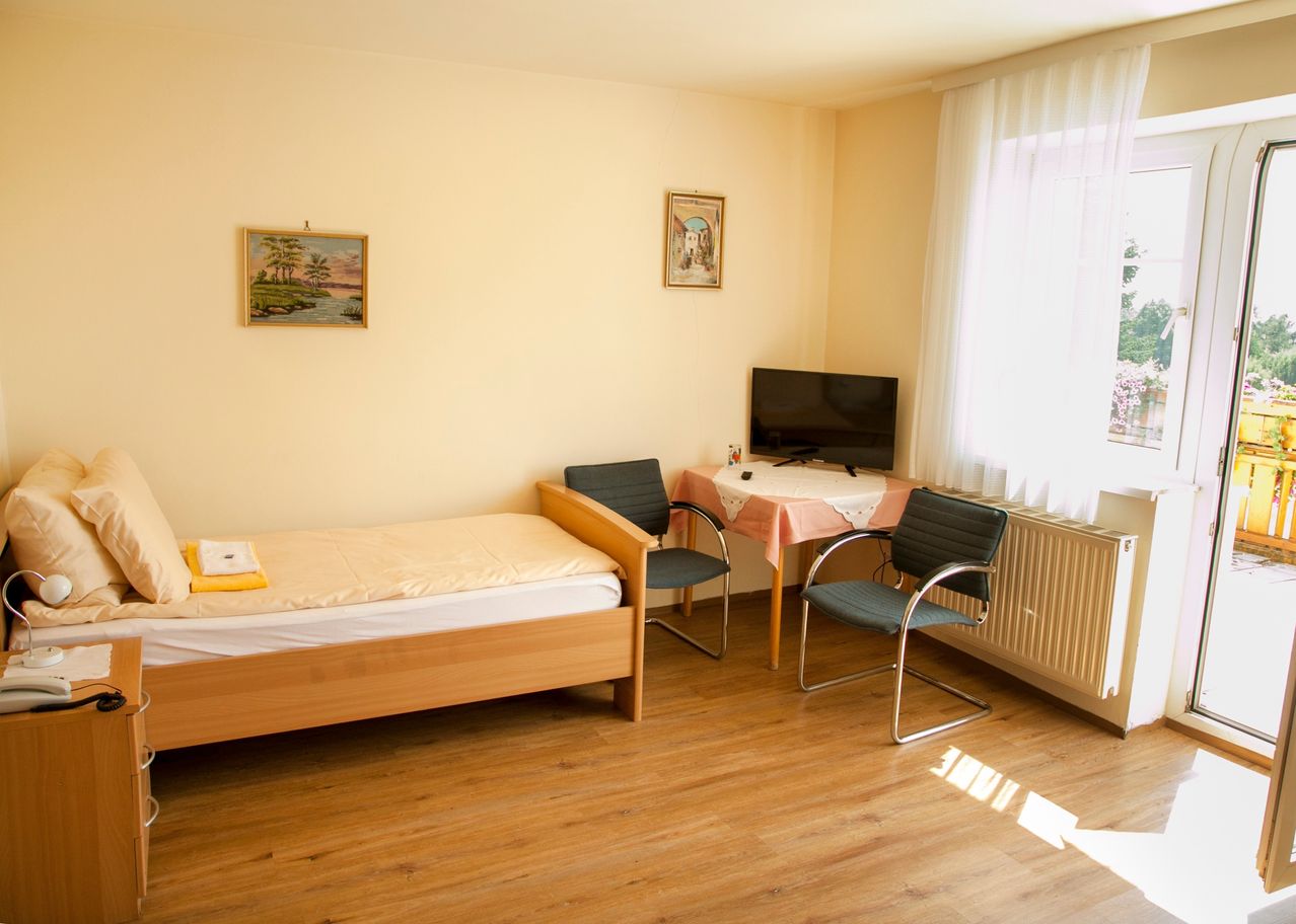 Ein einfaches Schlafzimmer mit einem Einzelbett, einem Schreibtisch mit Fernseher, zwei Stühlen und einem Heizkörper. Es gibt zwei Bilderrahmen an der Wand, und der Boden ist aus Holz.