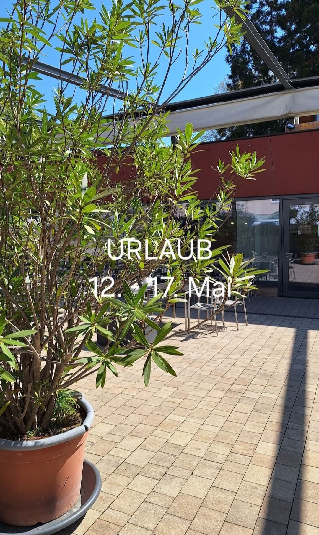 A green plant is outside a building with glass doors. The word 'URLAUB' is displayed above a date range. The date range is from May 12 to May 17.