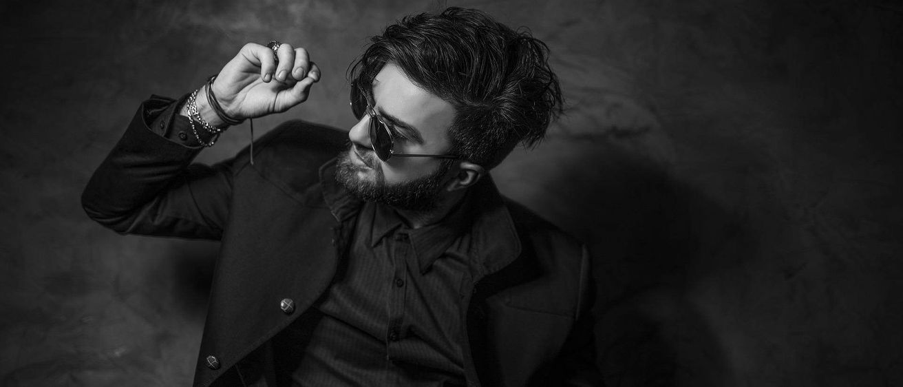 Ein Schwarz-Weiß-Porträt eines Mannes in einem Anzug, der einen Ring hält und eine Sonnenbrille trägt. Er wirkt nachdenklich, mit Bart und mehreren Armbändern am Handgelenk.