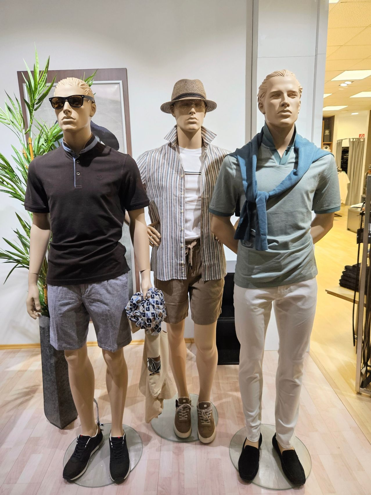 Drei Mannequins zeigen Herrenmode in einem Geschäft. Der Linke trägt ein schwarzes Polohemd mit Sonnenbrille. Der Mittlere trägt ein gestreiftes Hemd und Shorts. Der Rechte trägt ein blaues Hemd mit Schal und weißer Hose.