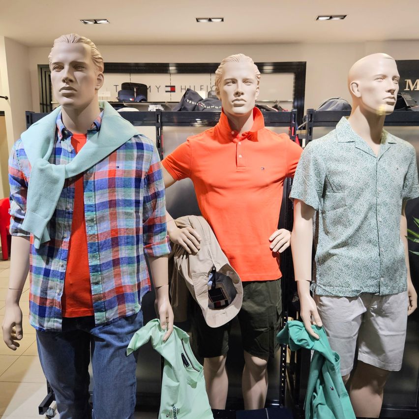 Drei Mannequins in einem Bekleidungsgeschäft zeigen verschiedene Outfits. Das linke trägt ein kariertes Hemd und einen Schal, das mittlere ein orangefarbenes Poloshirt und das rechte ein gemustertes Hemd und Shorts.