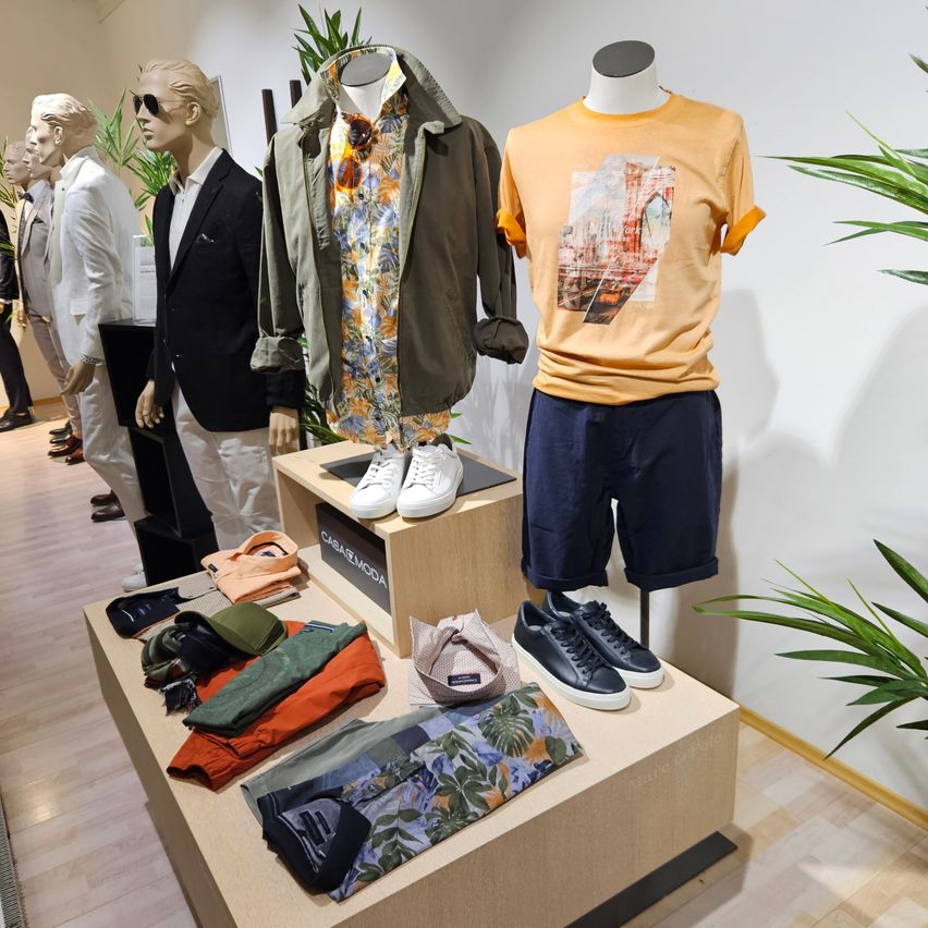 Eine Kleidungsausstellung mit Mannequins, die Anzüge, Blumenhemden und Sneaker tragen. Eine Schachtel mit dem Label Casamoda steht in der Mitte. Verschiedene Kleidungsstücke sind auf dem Tisch ausgestellt.