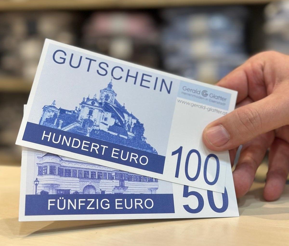 Eine Person hält zwei Geschenkgutscheine, einen für 100 Euro und einen für 50 Euro, mit einem blauen Gebäudedesign im Hintergrund.