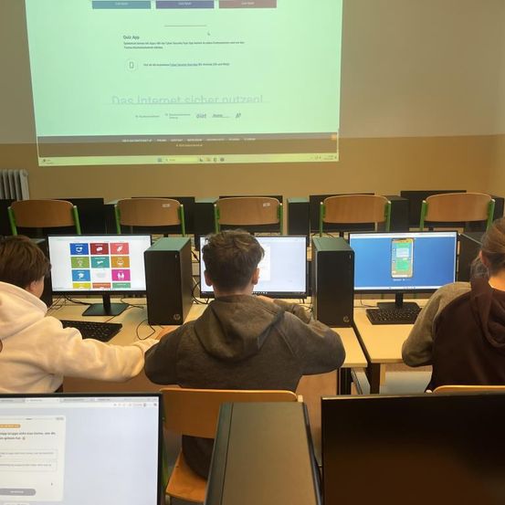Schüler sitzen in einem Klassenzimmer mit Computern, Stühlen und einer Projektionsleinwand. Die Leinwand zeigt eine Präsentation zur sicheren Internetnutzung.