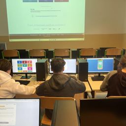 Schüler sitzen in einem Klassenzimmer mit Computern, Stühlen und einer Projektionsleinwand. Die Leinwand zeigt eine Präsentation zur sicheren Internetnutzung.