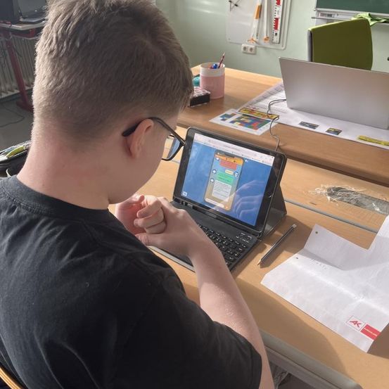 Ein Junge mit Brille benutzt ein Tablet, während er in einem Klassenzimmer sitzt. Seine Hände sind vor ihm verschränkt.