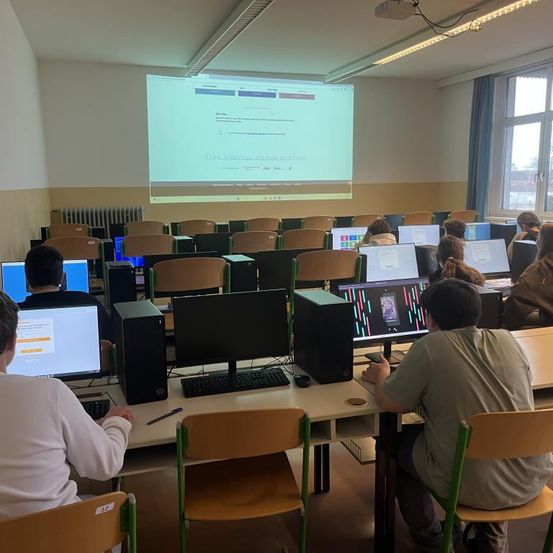 Schüler sitzen an Computern in einem Klassenzimmer. Eine Projektionsleinwand an der Wand zeigt eine Präsentation. Der Raum ist gut beleuchtet mit mehreren Fenstern und Deckenleuchten.