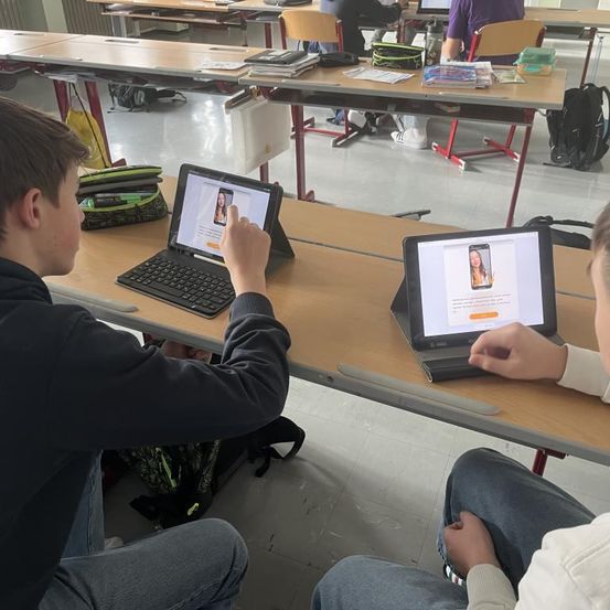 Zwei Schüler sitzen in einem Klassenzimmer und benutzen Tablets. Einer hat ein Bild einer Frau auf dem Bildschirm, während der andere es betrachtet. Ein Rucksack und eine Handtasche stehen neben ihnen.