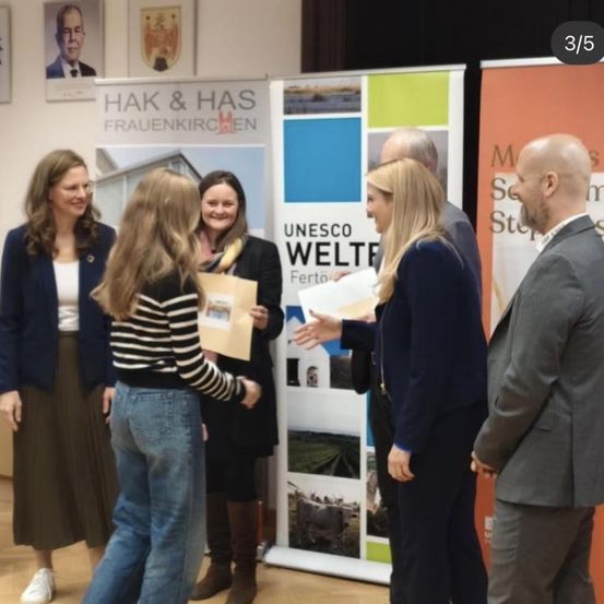 Fünf Personen stehen in einem Raum, zwei Frauen halten Papiere und unterhalten sich. Hinter ihnen sind Banner mit Bildern und Text, eines mit der Aufschrift 'UNESCO Welter'.