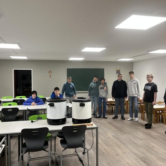 Mehrere Schüler stehen in einem Klassenzimmer um einen Tisch mit zwei großen Kochtöpfen. Ein Schüler sitzt auf einem Stuhl am Tisch. Andere stehen hinter dem Tisch.