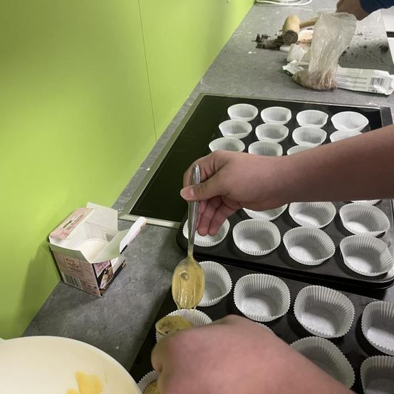 Eine Person füllt Cupcake-Förmchen mit Teig in einer Küche. Zwei Tabletts mit Cupcake-Förmchen stehen auf dem Tresen.
