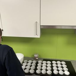 Eine Person in der Küche bereitet Cupcakes zu, mit Tabletts voller Cupcake-Förmchen auf dem Tresen und einer Schüssel in der Nähe. Weiße Schränke und eine grüne Wand bilden den Hintergrund.