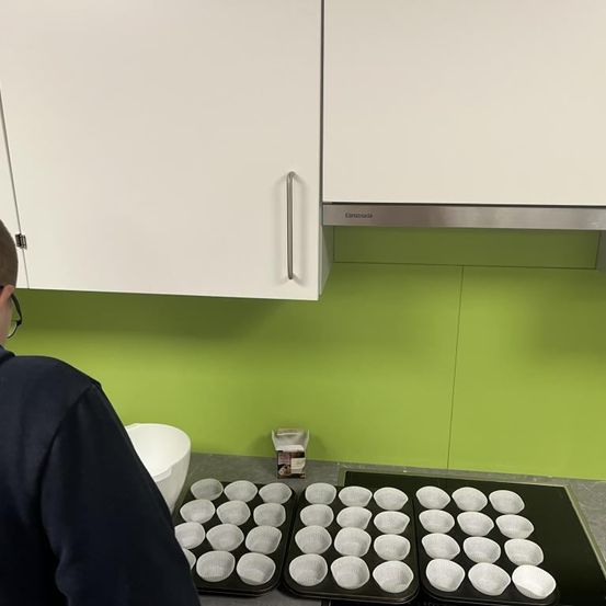 Eine Person in der Küche bereitet Cupcakes zu, mit Tabletts voller Cupcake-Förmchen auf dem Tresen und einer Schüssel in der Nähe. Weiße Schränke und eine grüne Wand bilden den Hintergrund.