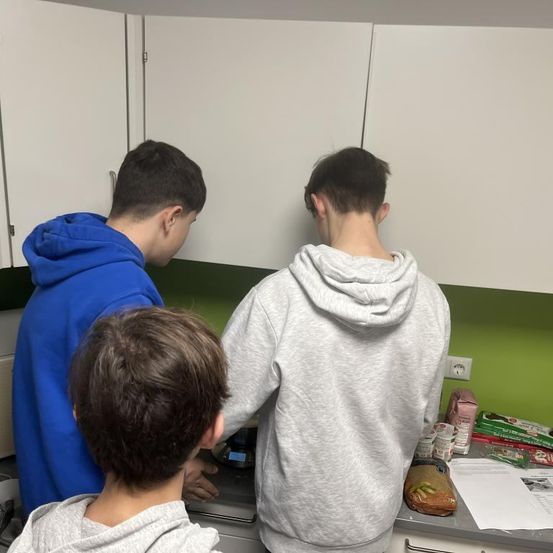 Drei Jungen befinden sich in einer Küche und bereiten Essen zu. Der Junge in der Mitte schaut auf die Waage. Der Junge auf der rechten Seite steht und schaut auf den Jungen in der Mitte. Der Junge auf der linken Seite beugt sich vor und schaut auf etwas.
