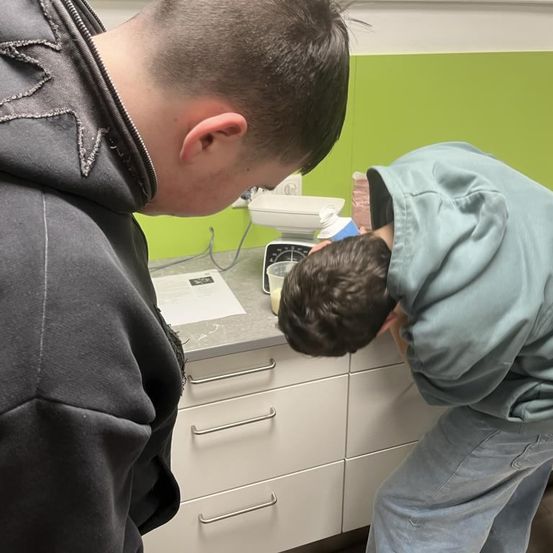 Zwei Jungen sind in einer Küche; einer beugt sich über ein Waschbecken, während der andere zusieht. Der Junge am Waschbecken hält einen Becher in der Hand.
