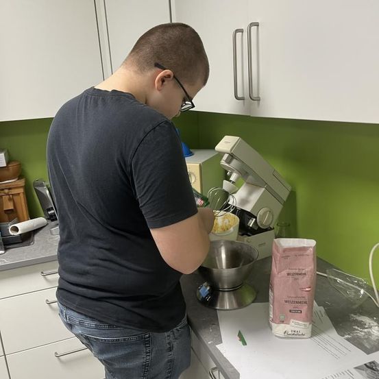 Ein Mann mit Brille steht in einer Küche und benutzt einen Standmixer, um Zutaten in einer Schüssel zu vermischen. Der Arbeitstisch ist mit verschiedenen Küchenutensilien übersät.