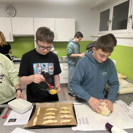 Mehrere junge Jungen bereiten Essen in einer Küche zu. Ein Junge mit Brille trägt Eigelb auf den Teig auf, während ein anderer den Teig ausrollt. Eine Ablage mit ausgerolltem Teig steht auf dem Tresen.