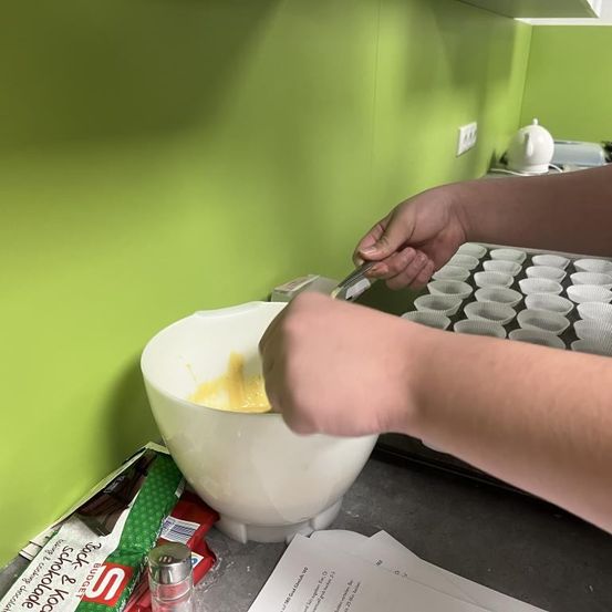 Eine Person mischt Zutaten in einer Schüssel in einer Küche. Die Schüssel steht auf einem Arbeitstisch. Cupcakes-Backformen sind im Hintergrund zu sehen.