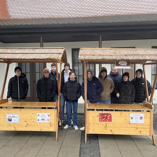 Eine Gruppe junger Leute steht vor zwei hölzernen Ständen mit Strohdächern und verkauft Essen. Ein Stand verkauft Muffins und der andere verkauft Lebkuchen.