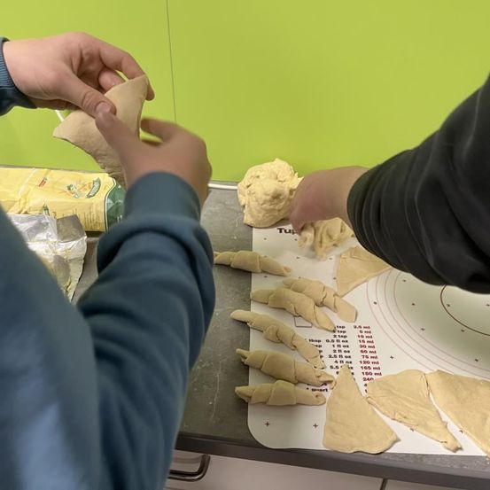 Zwei Personen arbeiten an Teig, möglicherweise Brot oder Gebäck. Einer rollt und schneidet den Teig, während der andere ein Stück hält. Sie befinden sich in einer Küche mit einer grünen Wand und einem Messmatte.