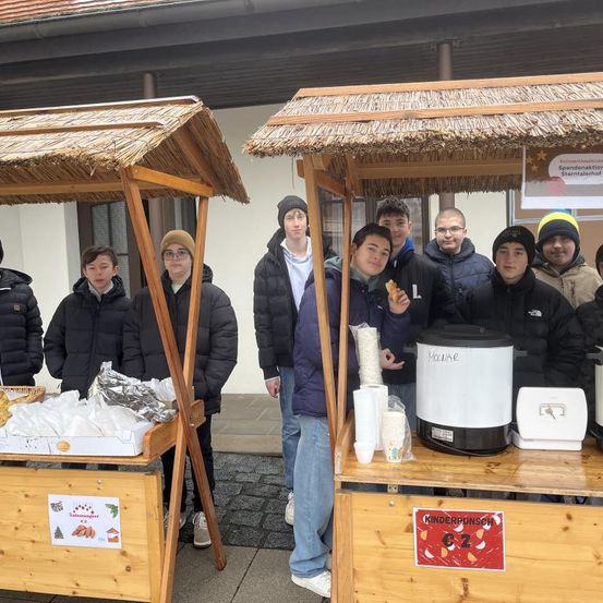 Eine Gruppe von Jungen steht hinter einem hölzernen Stand mit Essen auf einem Tisch. Ein weiterer Stand mit Kaffee und heißer Schokolade ist in der Nähe. Sie tragen alle Winterkleidung.