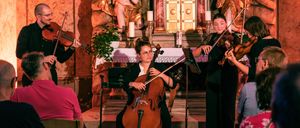 Ein klassisches Musikensemble spielt in einer Kirche, mit Musikern auf Cello, Geige und Bratsche. Dahinter befindet sich ein Altar mit Statuen und Kerzen, der eine dramatische Kulisse schafft. Das Publikum sitzt vorne und beobachtet aufmerksam.