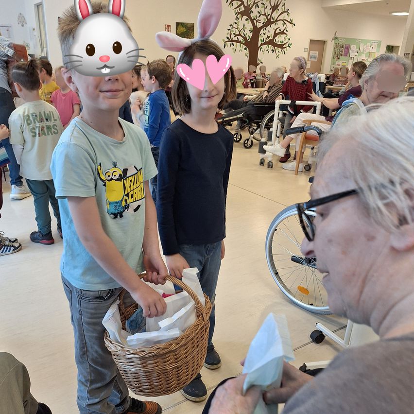 Zwei Kinder mit Hasenöhrchen halten einen Korb mit Geschenken für eine ältere Frau in einem Rollstuhl. Dahinter stehen andere Kinder, einige sitzen in Rollstühlen, andere stehen. An der Wand befindet sich ein Baumwandbild.