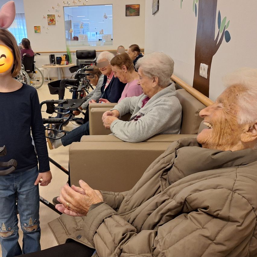 Ein junges Mädchen mit Hasenohren lächelt und steht neben einer älteren Frau in einem Stuhl mit einem lächelnden Gesicht. Hinter ihnen sitzt eine Gruppe älterer Frauen auf einer Couch in einem Raum mit einer Wand, die einen Baum und ein Fenster zeigt.