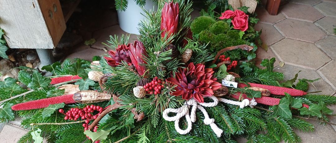 Ein festlicher Kranz mit roten Blumen, grünen Blättern und einer weißen Schleife liegt auf einem gefliesten Boden, umgeben von anderen Kränzen.