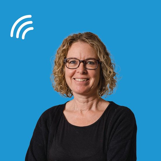 Eine lächelnde Frau mit Brille und schwarzem Oberteil steht vor einem blauen Hintergrund mit einem weißen Symbol.