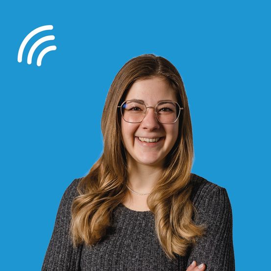 Eine Frau mit Brille und langen, welligen Haaren lächelt und posiert für ein Foto vor blauem Hintergrund.