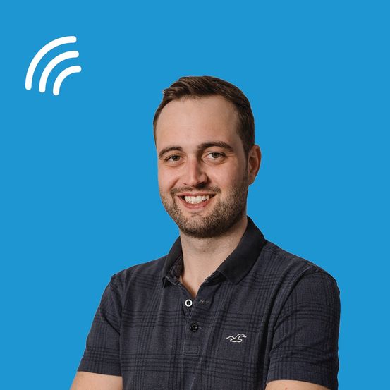 Ein Mann mit Bart in einem schwarzen Poloshirt lächelt und posiert für ein Foto mit blauem Hintergrund und weißem WLAN-Symbol.