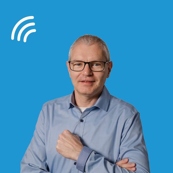 Ein Mann mit Brille steht mit verschränkten Armen. Er trägt ein blaues Hemd und schaut direkt in die Kamera. Er hat ein weißes WLAN-Symbol in der oberen linken Ecke.