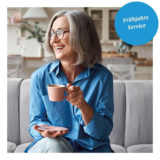 Eine ältere Frau sitzt auf einem Sofa und hält eine rosa Tasse mit Untertasse, lächelt. Der Hintergrund zeigt eine Küche mit weißen Möbeln und grünen Pflanzen. Ein blauer Kreis mit dem Text 'Fruhjahrs Service' ist sichtbar.