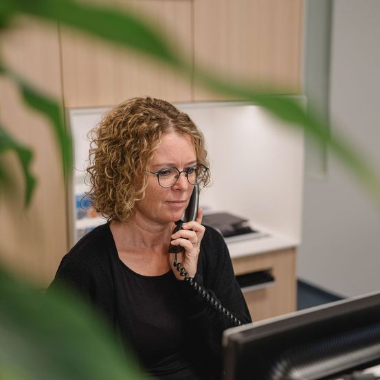 Eine Frau mit lockigem Haar und Brille sitzt an einem Schreibtisch im Büro und hält ein Telefon an ihr Ohr. Sie lächelt und scheint ein Gespräch zu führen.