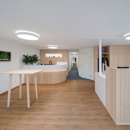 Ein offener Empfangsbereich mit einem Schreibtisch und einer Frau, die daran sitzt. Holzböden, weiße Wände und ein modernes Design.