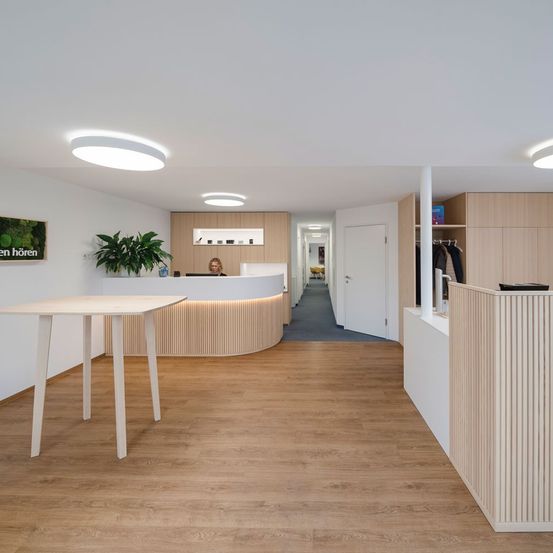 Ein offener Empfangsbereich mit einem Schreibtisch und einer Frau, die daran sitzt. Holzböden, weiße Wände und ein modernes Design.