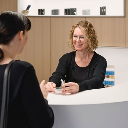Zwei Frauen unterhalten sich in einem modernen, hellen Raum. Eine Frau lächelt, hält einen Stift, während die andere Frau aufmerksam zuhört. Hinter ihnen sind Holzschränke mit Regalen an der Wand montiert.