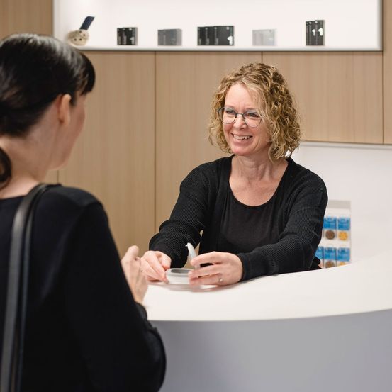 Zwei Frauen unterhalten sich in einem modernen, hellen Raum. Eine Frau lächelt, hält einen Stift, während die andere Frau aufmerksam zuhört. Hinter ihnen sind Holzschränke mit Regalen an der Wand montiert.