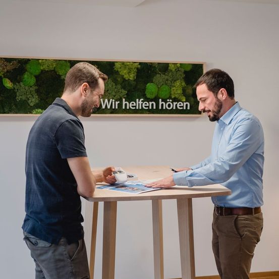 Zwei Männer stehen vor einem Holztisch. Einer sieht in ein Büchlein, während der andere einen Stift hält und auf ein Blatt Papier schaut. Hinter ihnen ist eine weiße Wand mit einer großen grünen Pflanze und einem weißen Schild.