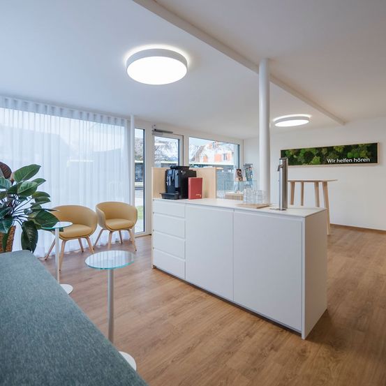 Ein modernes, sauberes Zimmer mit weißen Schränken, einer Arbeitsplatte mit Kaffeemaschine und einem Holzboden. Zwei gelbe Stühle und eine Pflanze stehen neben einem Fenster mit weißen Jalousien. Ein Poster an der Wand sagt: 'Wir helfen Ihnen zu hören.'