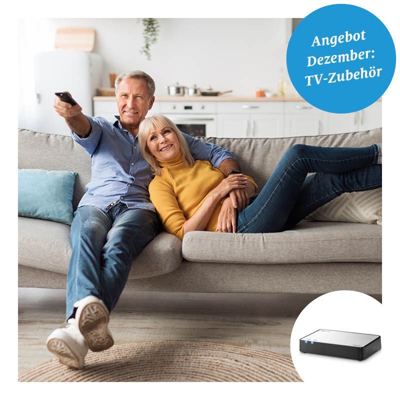 Ein älteres Paar schaut fern auf einer Couch im Wohnzimmer. Der Mann hält eine Fernbedienung und die Frau lächelt in die Kamera. In der unteren rechten Ecke befindet sich ein TV-Zubehör.