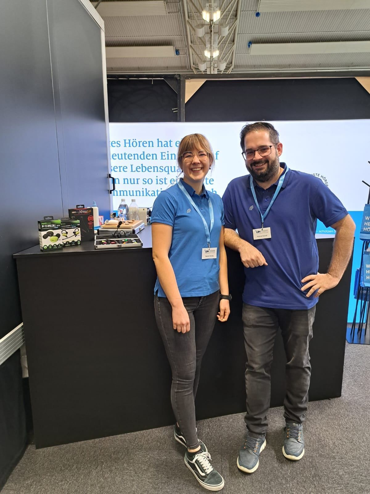 Two people in blue shirts stand behind a counter, smiling. A screen displays text in German. Various items are on the counter.