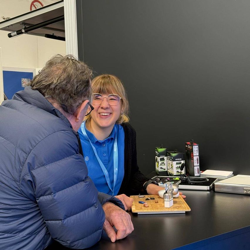 An older man wearing glasses is looking at a smiling woman in front of him. They are sitting at a desk with various items, including a box, a laptop, and a game board. The background shows a gray wall.