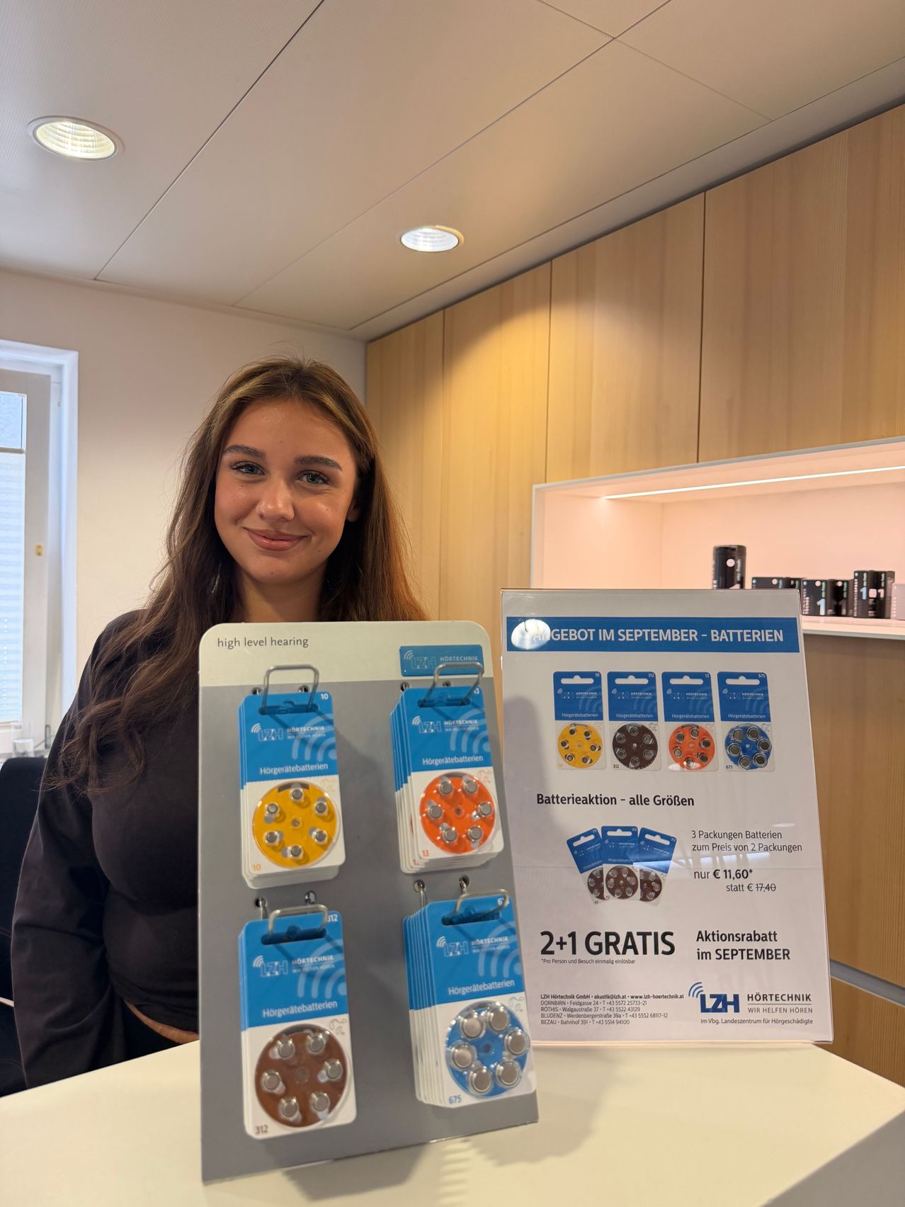 A woman stands next to a display of hearing aid batteries, smiling. The display features a promotional offer for September with three packs of batteries for the price of two.